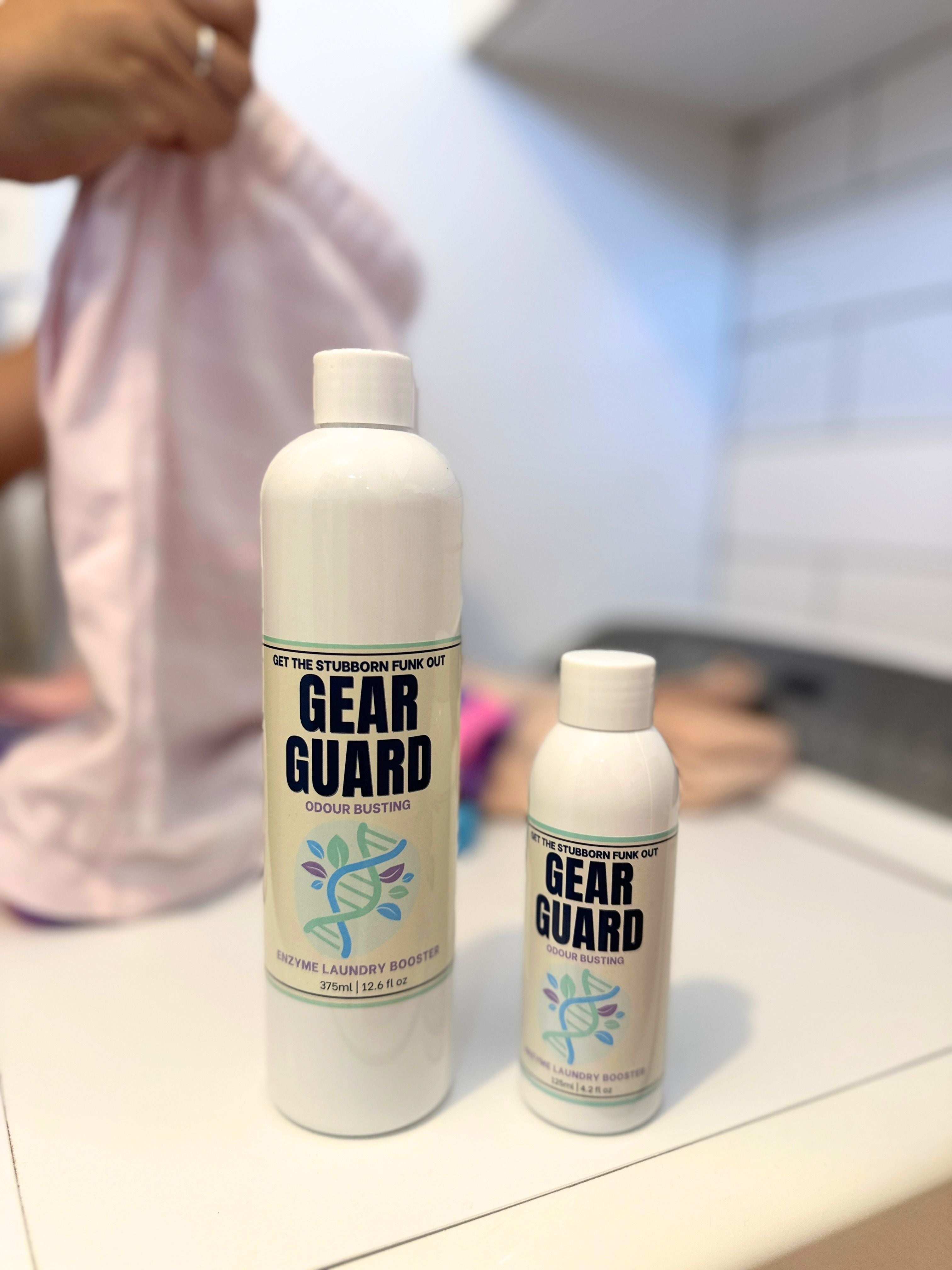 Two bottles of Gear Guard laundry booster on a washing machine with someone folding laundry blurred behind