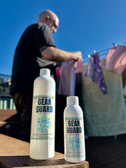 Two bottles of Gear Guard laundry booster on a table with a person hanging clothes in the background.