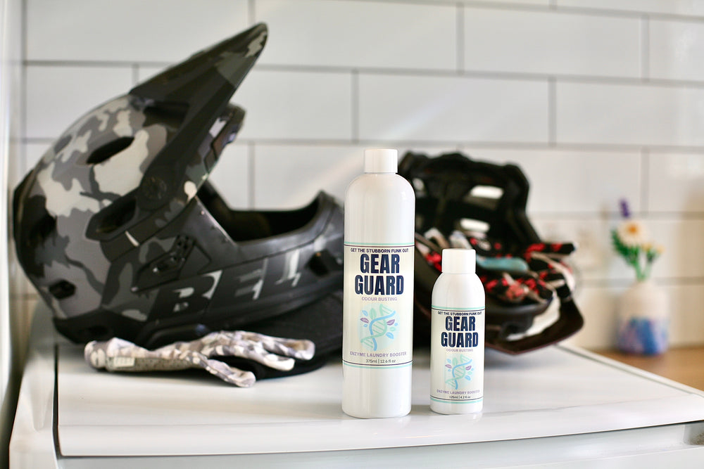 Two 'Gear Guard' bottles on a washing machine with Mountain Biking helmets in the background.