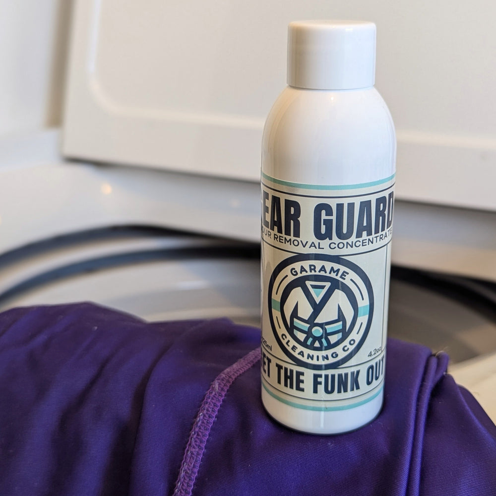 A single bottle of Gear Guard sits on top of a purple rash guard. The rash guard is draped over the edge of a washing machine.