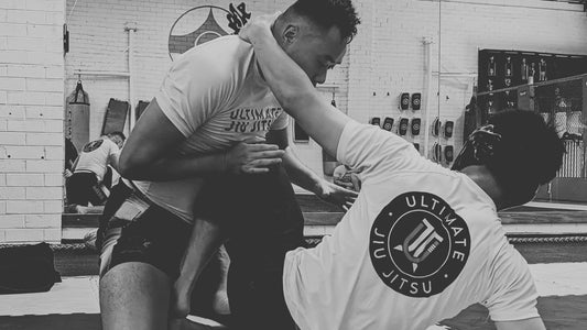 A black and white photograph captures a dynamic Jiu-Jitsu training moment, with both practitioners in white rash guards and black shorts, one sitting down propped up on their elbow. 