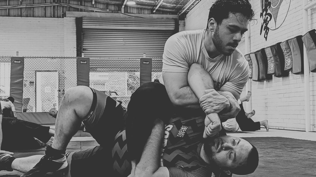 This image shows two individuals engaged in a Brazilian Jiu-Jitsu training session on a black mat. 