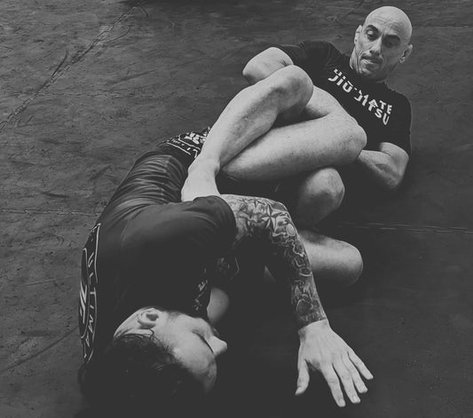 Jiu-Jitsu students practice submission techniques on the mat, with a focused athlete in a black rash guard securing a leg lock against another student in a blue rash guard.