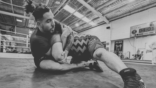 Two athletes engage in a Brazilian Jiu-Jitsu drill on black mats in a gym, with the person in a blue rash guard executing a top control position over the defender in patterned red and black shorts.