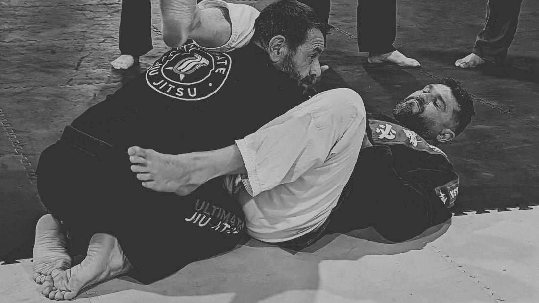 A Jiu-Jitsu instructor in a black gi oversees a demonstration as students, wearing gis of various colors, watch attentively, with two practitioners on the mat in a grappling position.