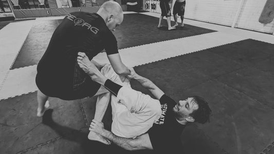 A black and white photo captures an intense Brazilian Jiu-Jitsu training session with one practitioner in a black rash guard and white pants on his back executing a guard while the other in a black rash guard and shorts works to pass.