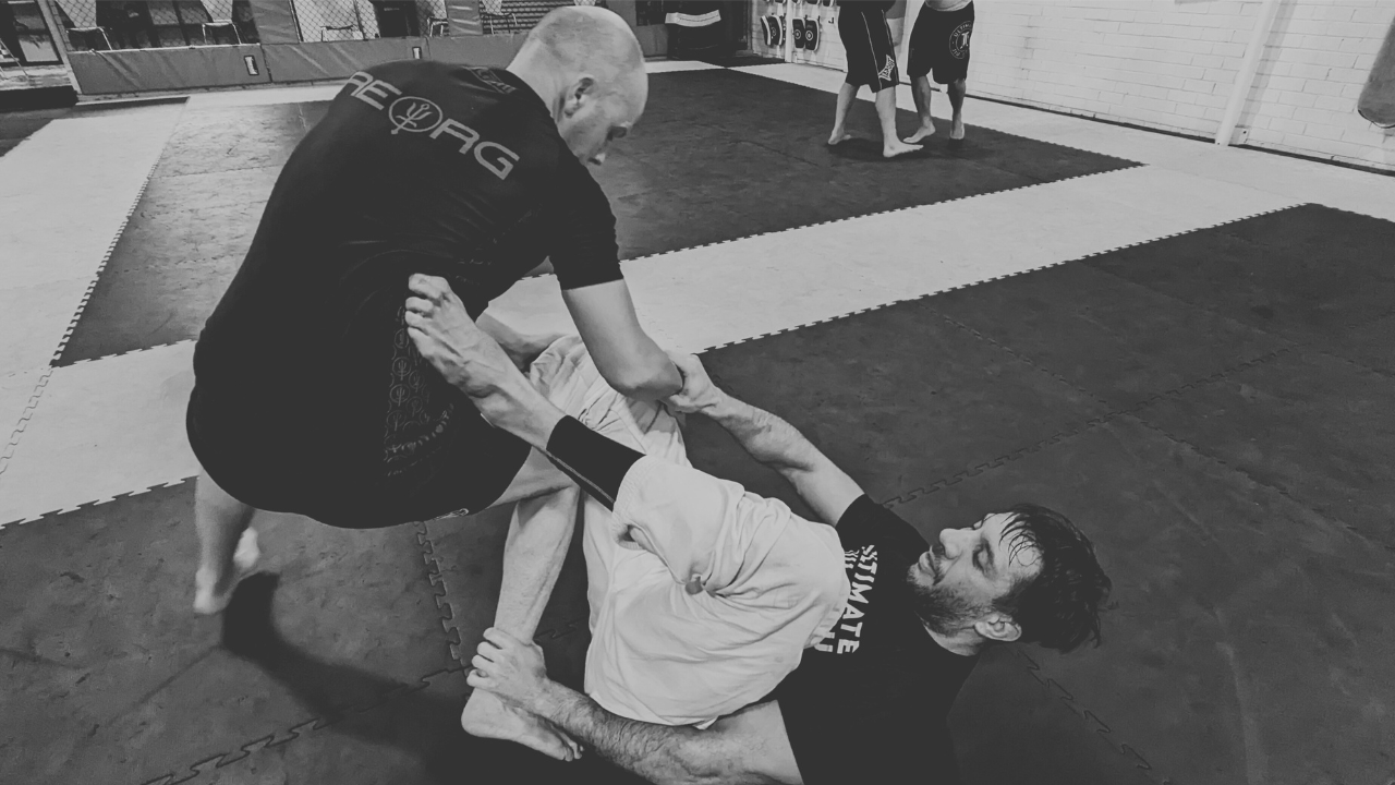 A monochrome image of two people locked in a brazillian jiujitsu match. One person is on their back, the other in a semi squat position. The person on the ground is holding the opponents wrist with his right hand, and the ankle with the other.