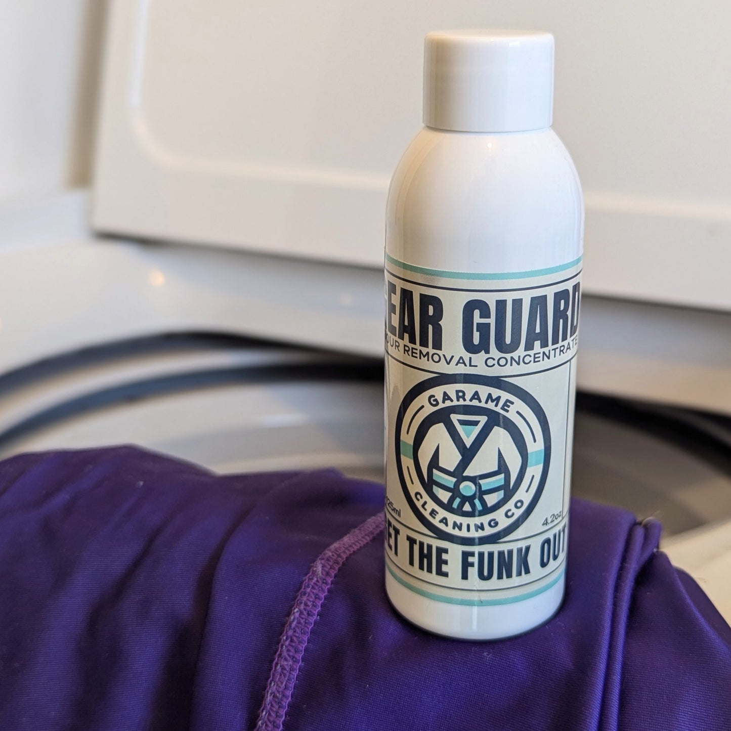 A single bottle of Gear Guard sits on top of a purple rash guard. The rash guard is draped over the edge of a washing machine. 