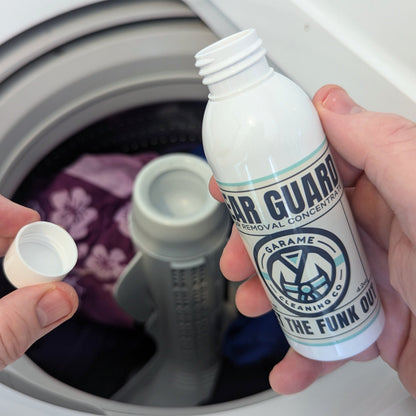 Ther eis a bottle of Gear Guard in one hand, and the cap in the other, poised over the top of a washing machine with training gear in it. This shows that you only need one capful of Gear Guard in each load of washing. 