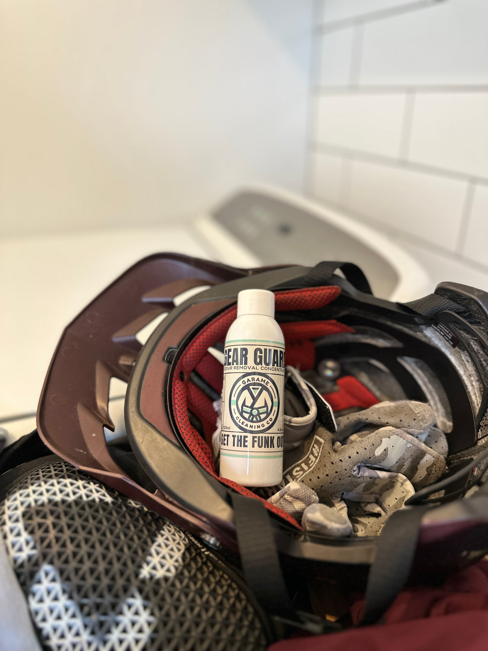 Bottle of Gear Guard inside a helmet with riding gloves, sitting next to a washing machine.