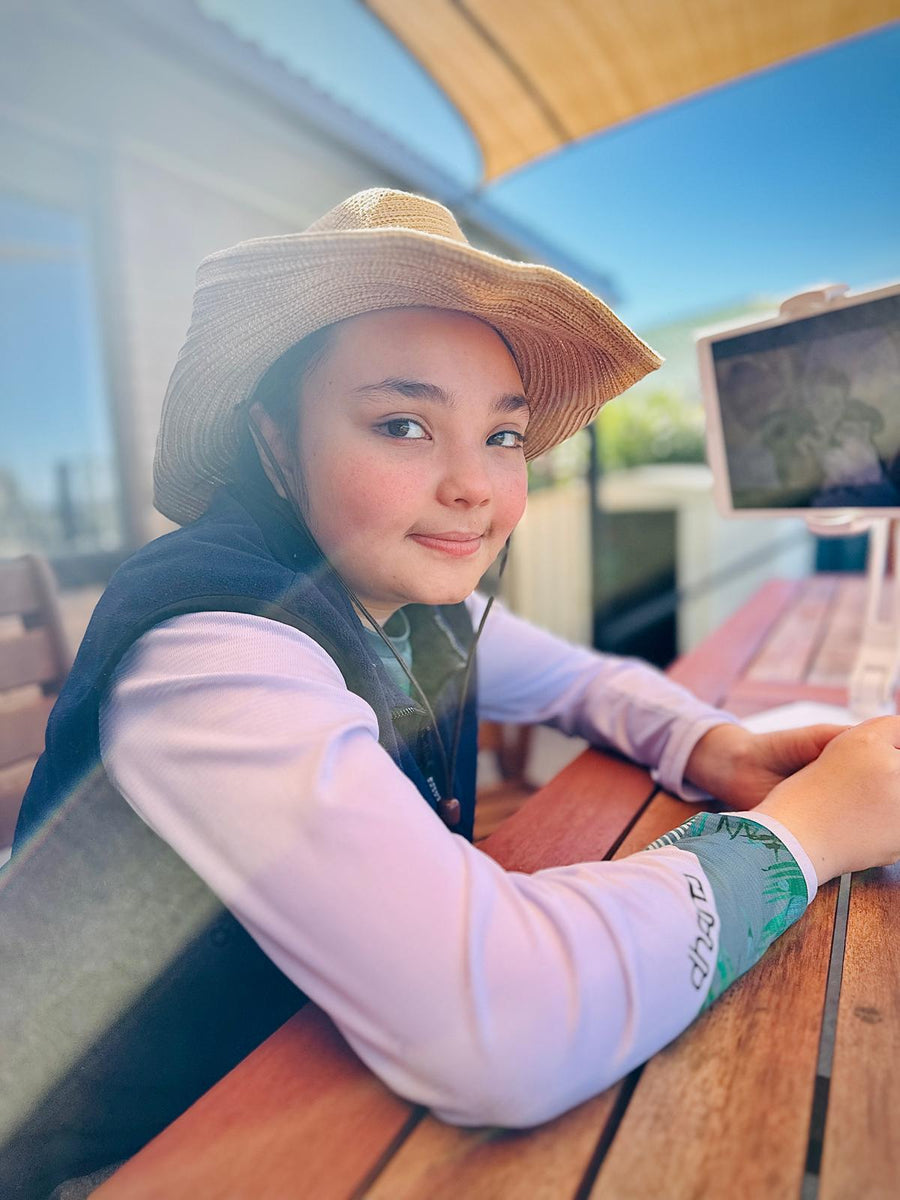 Kenzie smiling at the camera under a shade sail at a wooden table, wearing a straw hat, with her iPad showing The Little Mermaid beside her