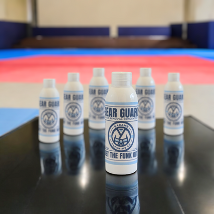 Six bottles of Gear Guard odour removal concentrate arranged neatly on a black table or bench in a gym, emphasizing the quantity and bulk purchase option. Each bottle features the same branding and design, with the company's logo and the 'GET THE FUNK OUT' tagline visible.